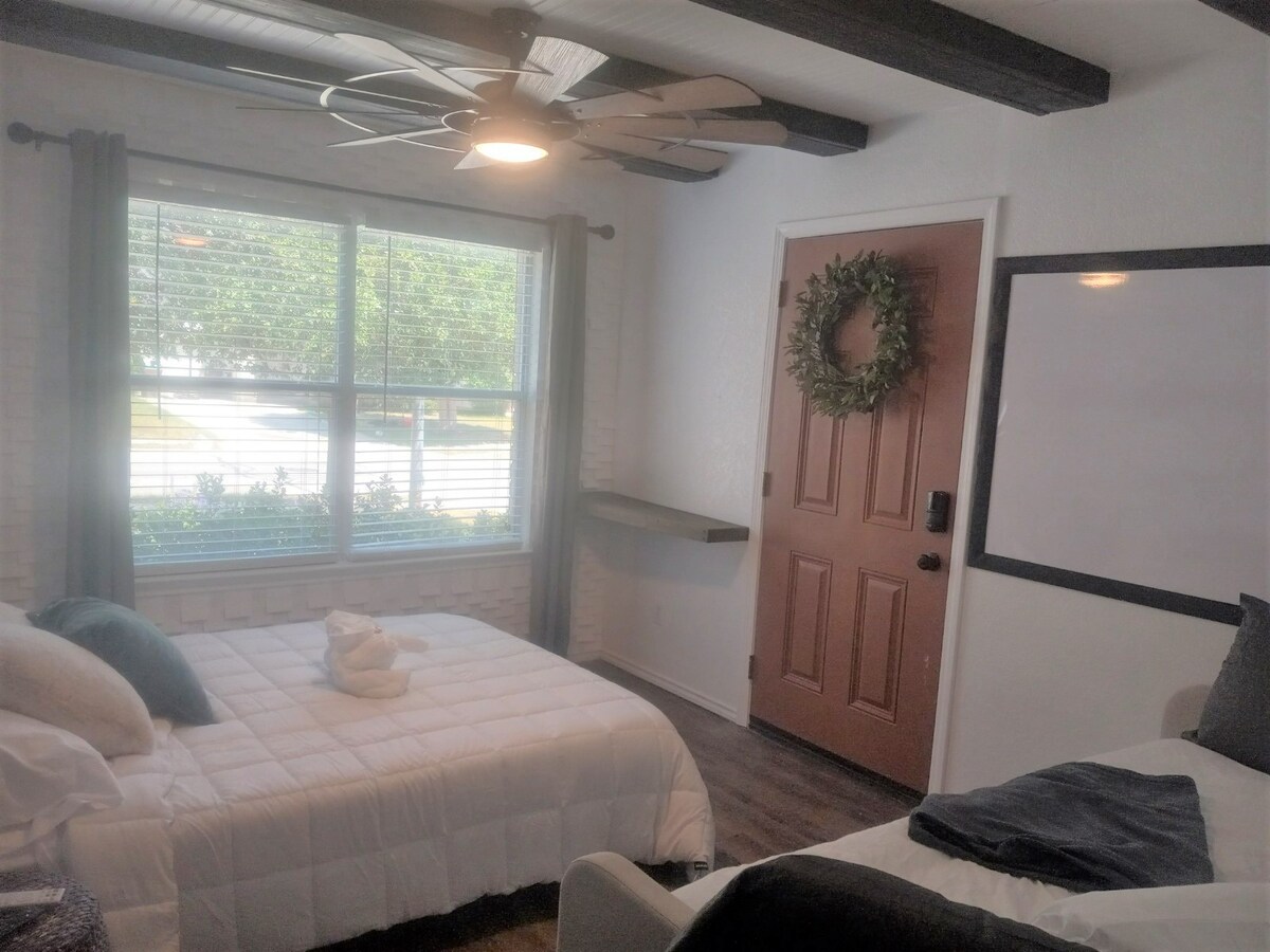 A well-arranged room features a queen bed and a twin daybed with a trundle, both covered with soft bedding. A ceiling fan hangs overhead, and a large window allows natural light to enter, providing views of the outside greenery. The door is adorned with a wreath.