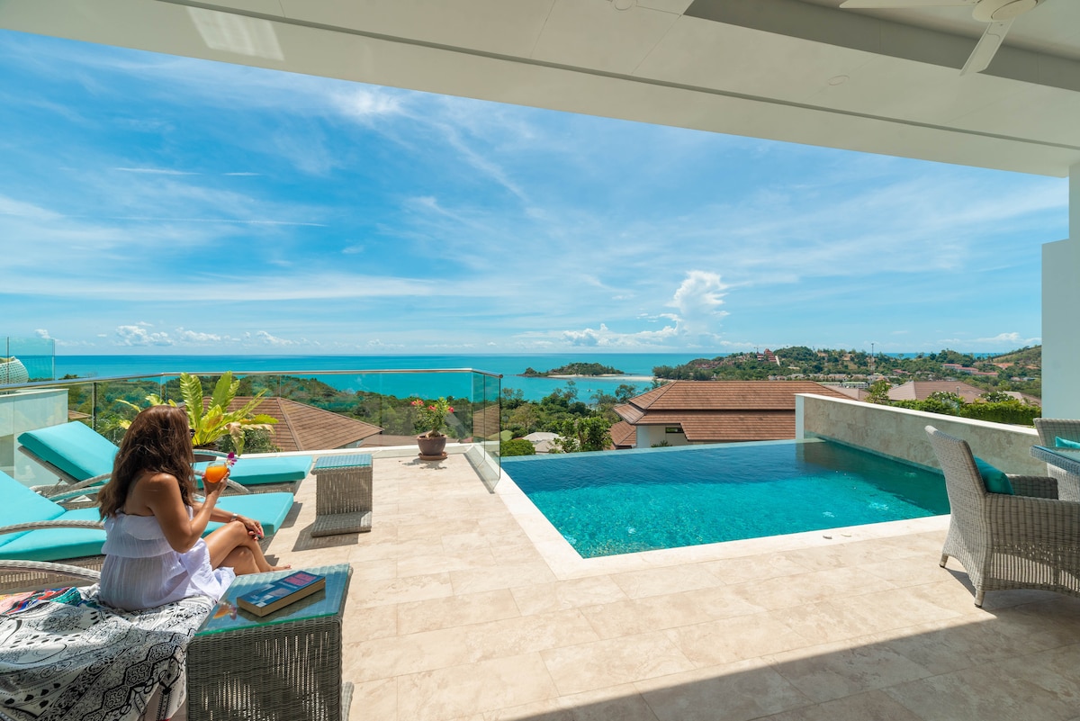 An outdoor terrace features a private infinity pool overlooking scenic views of the water. Comfortable seating areas are placed nearby, with a guest enjoying a drink, complemented by lush greenery and the expansive sky above.