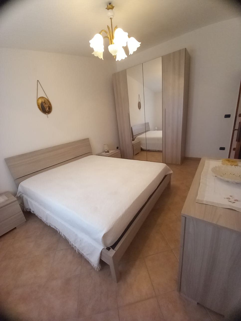 A simple bedroom is presented with a spacious bed covered in a light-colored blanket. A modern wardrobe with mirrored doors provides reflection. A light fixture with gentle illumination hangs from the ceiling. Neutral tones are paired with tiled flooring, contributing to a calming atmosphere.