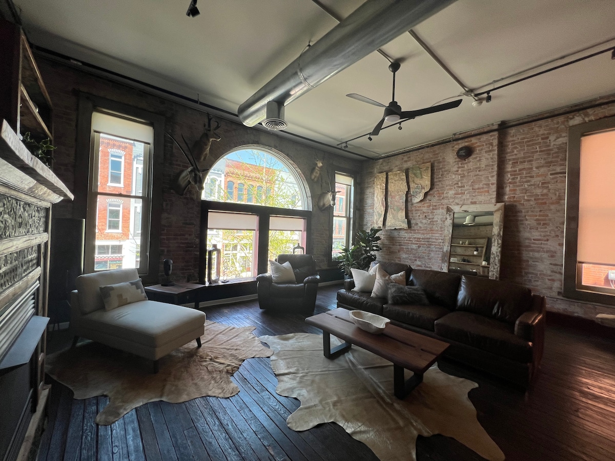 The spacious living area features large windows overlooking C-Street, allowing natural light to illuminate the exposed brick walls. A comfortable leather sofa and a light-colored chaise lounge are positioned on a textured area rug, creating an inviting space for relaxation.