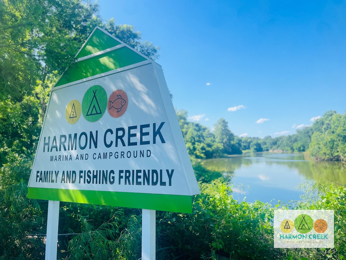 A welcoming sign for Harmon Creek Marina and Campground is positioned near a calm body of water, framed by lush greenery. The sign features symbols indicating family and fishing-friendly amenities, with blue skies and scattered clouds visible in the background.