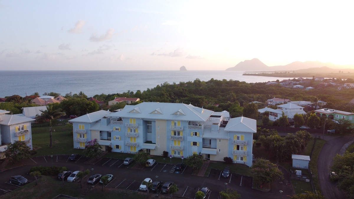 An aerial view captures a residential building surrounded by lush greenery, with the ocean visible in the background. The structure features multiple balconies with yellow shutters, and a parking area is situated in the foreground. Scenic coastal hills add to the tranquil setting.