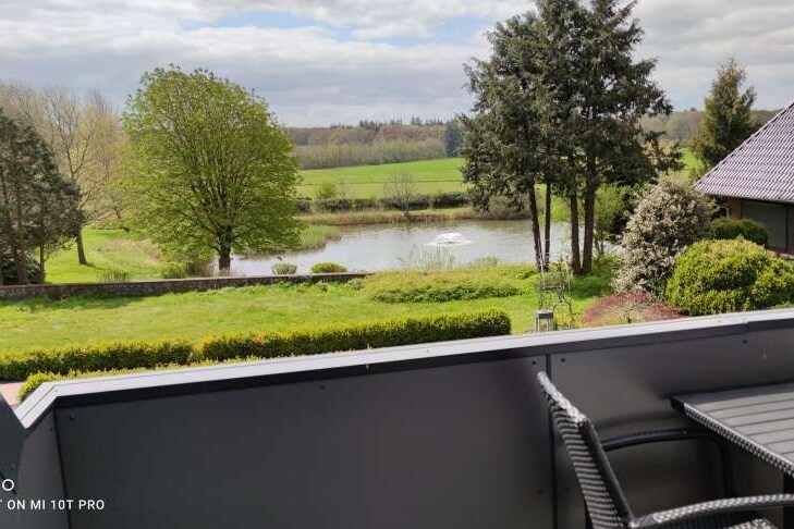 A balcony view captures a serene landscape featuring a private lake surrounded by lush greenery. An assortment of trees and shrubs frames the scene, while a well-maintained lawn extends towards the water's edge, offering a peaceful atmosphere.
