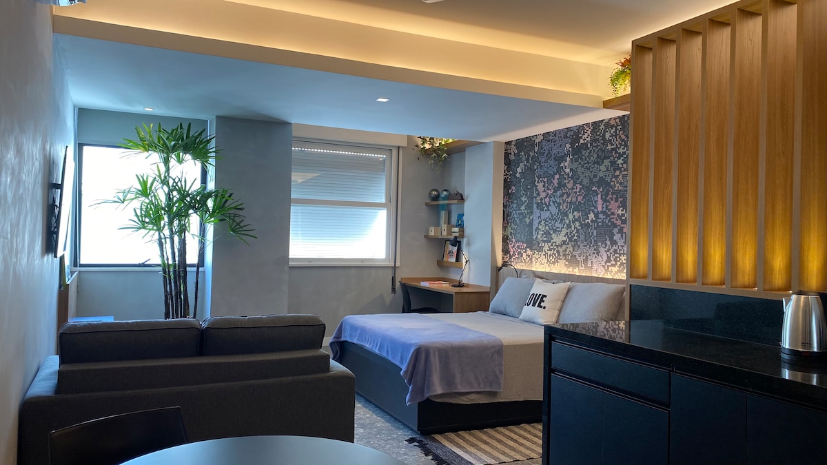 A contemporary loft space features a cozy sleeping area with a spacious bed, complemented by soft bedding and a decorative wall. A small sofa is placed nearby, alongside a dining table. Natural light enters through the window, with greenery adding a touch of freshness.