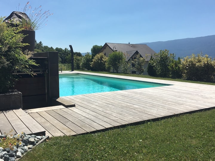 A tranquil pool area is presented, featuring a rectangular swimming pool surrounded by a wooden deck. Lush greenery and shrubbery border the space, with mountains visible in the distance under a clear blue sky.