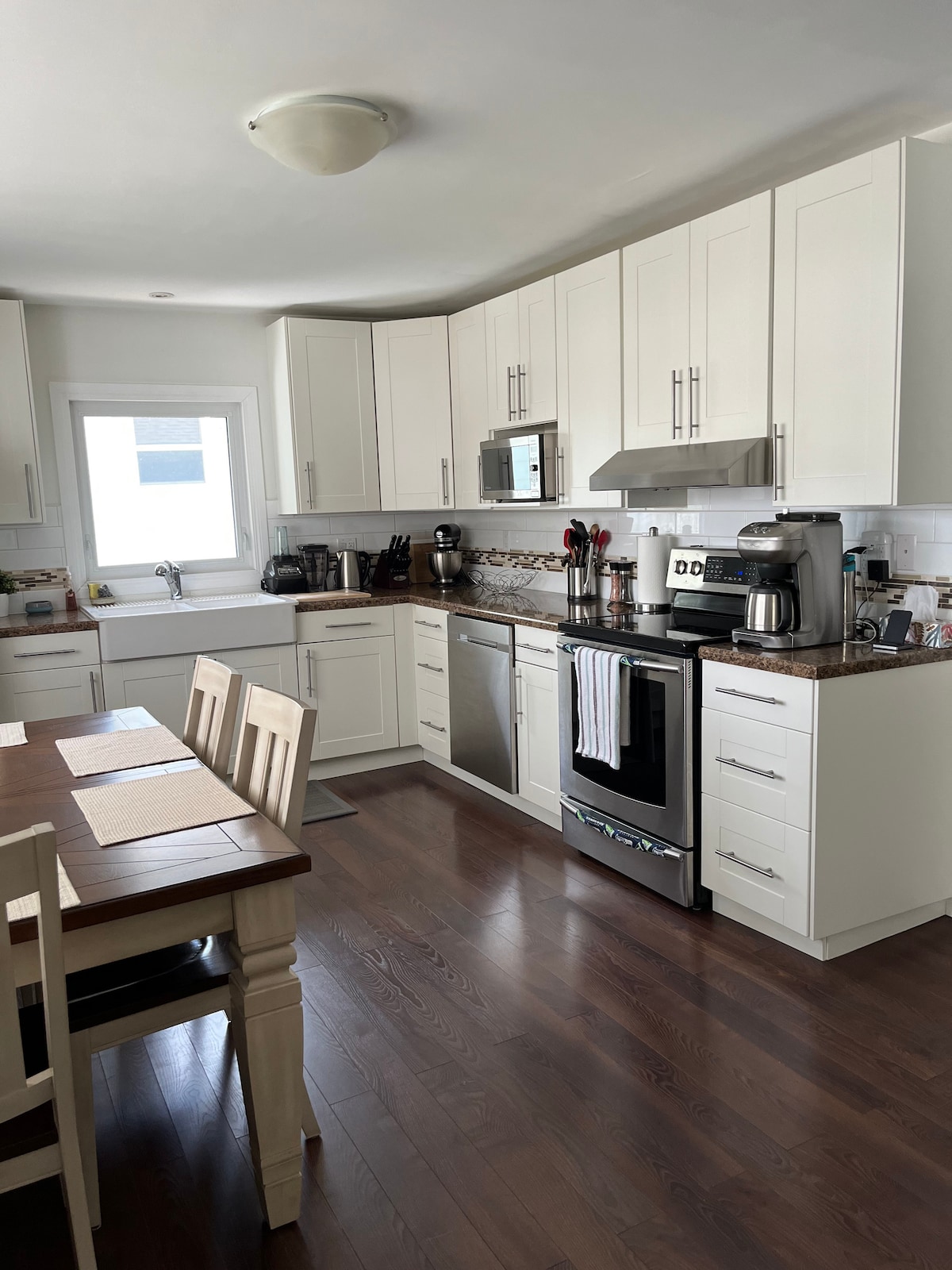 A modern kitchen features white cabinetry and stainless steel appliances. The countertop is adorned with kitchen essentials, and a dining table with light wooden chairs is positioned nearby. Natural light enters through a window above the sink, illuminating the space.