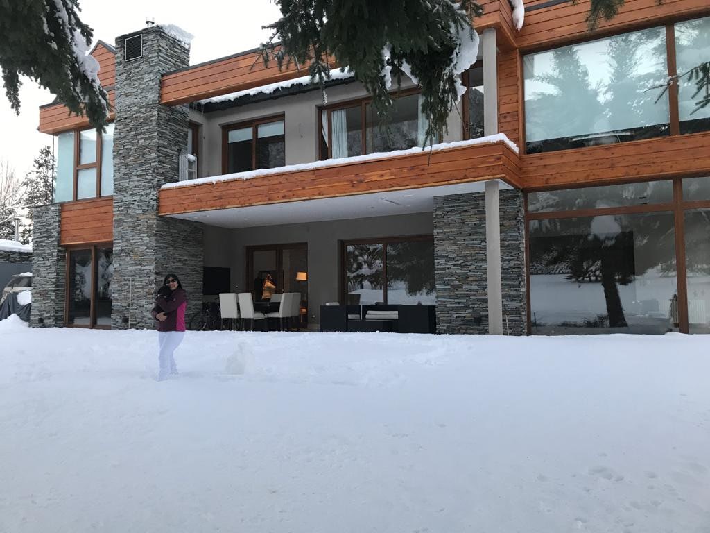 The modern facade of a two-story home is visible, featuring a mix of stone and wood materials. Snow covers the ground, and large glass windows reflect the snowy landscape. An outdoor seating area is seen on the porch, ready for relaxation.