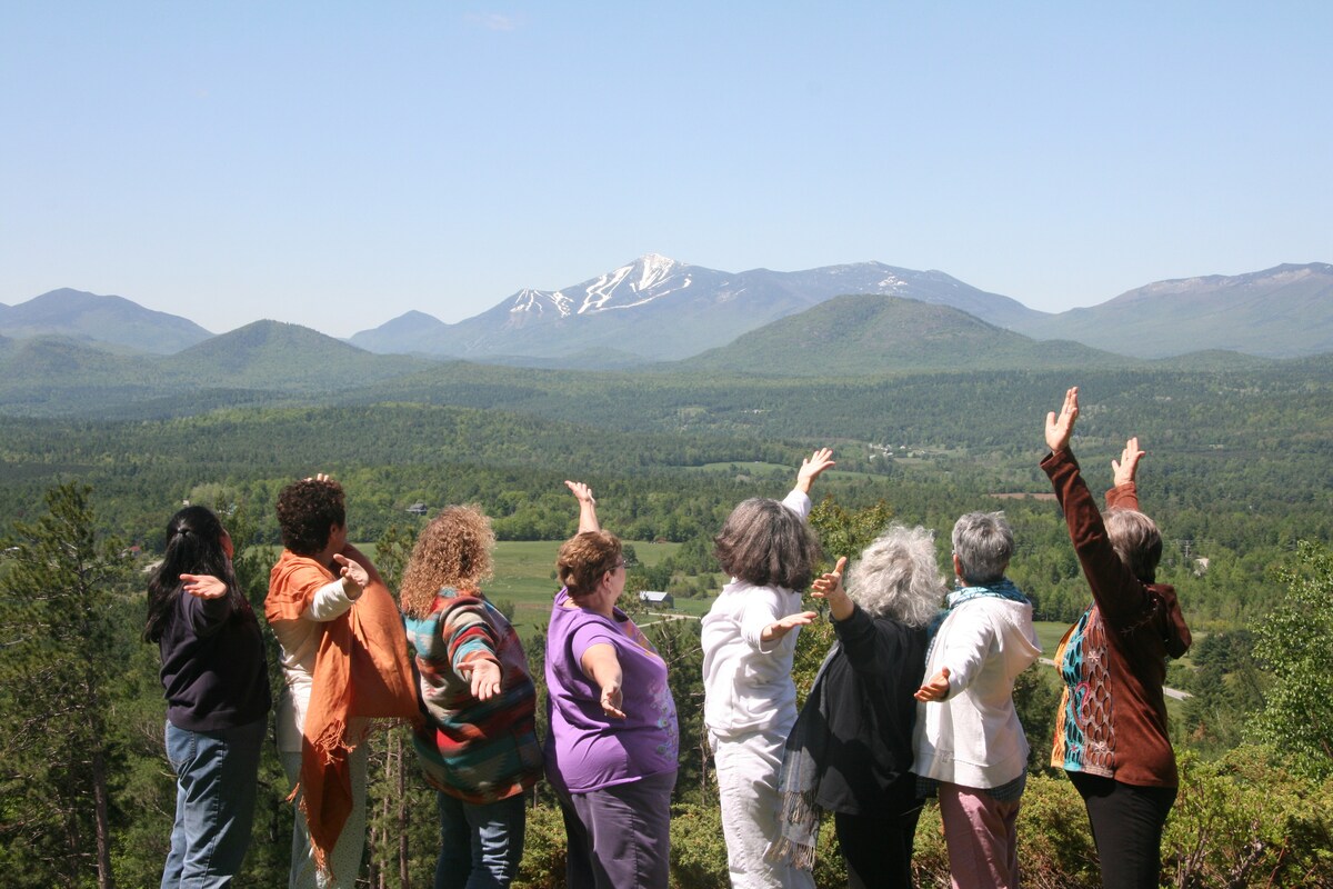 A group of eight individuals stands with arms extended towards a panoramic view of lush green hills and distant, snow-capped mountains. The scene captures a clear blue sky above, enhancing the vibrant landscape.