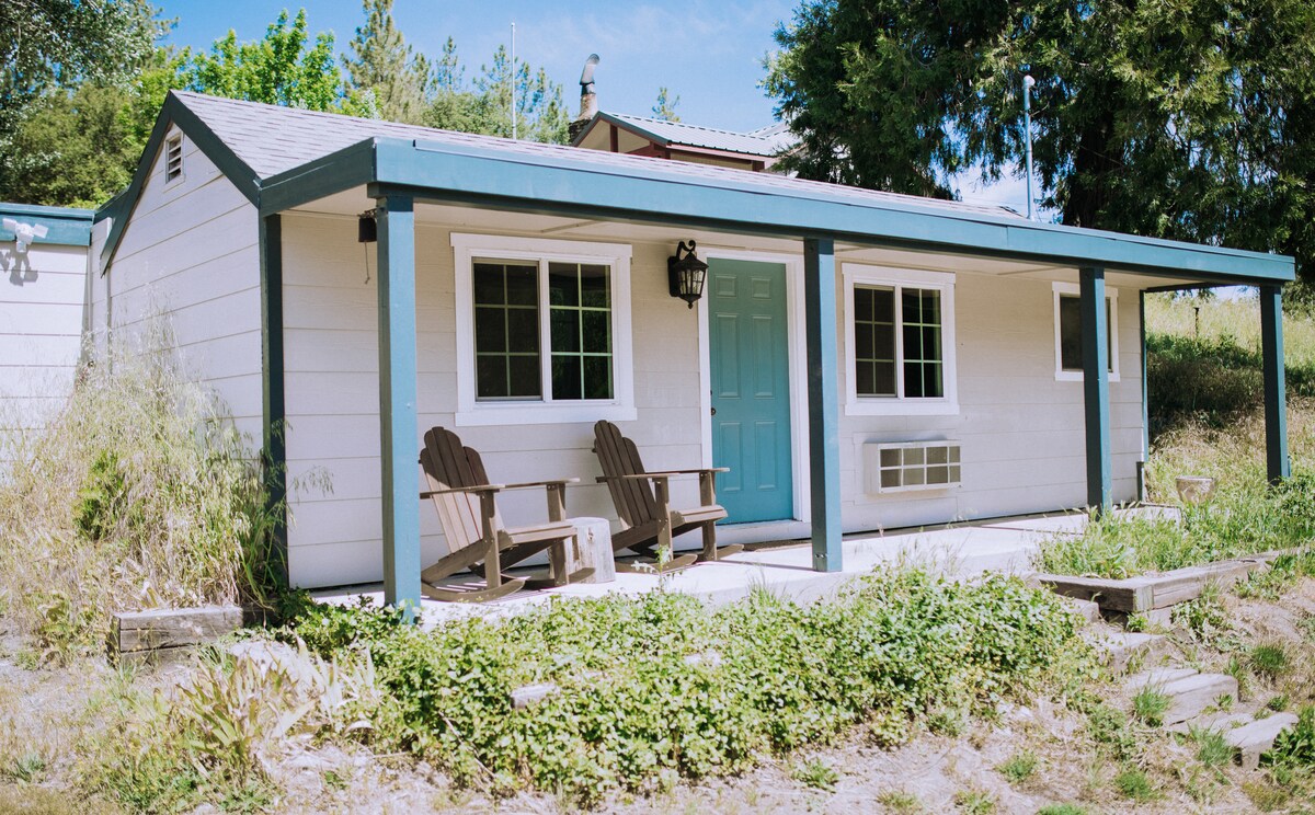The exterior of the cabin features a welcoming porch with two classic wooden chairs positioned to face the natural surroundings. A bright blue door stands out against the light-colored siding, while multiple windows allow for ample natural light, framed by greenery.