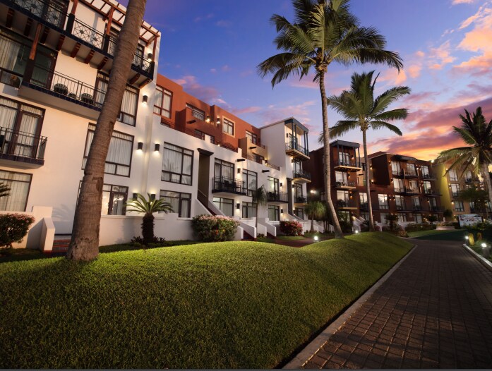 A modern resort building is viewed from the landscaped grounds, featuring multiple balconies and large windows. Palm trees frame the scene, while the path is bordered by lush greenery, illuminated by soft lighting. The sky transitions to dusk with warm colors.