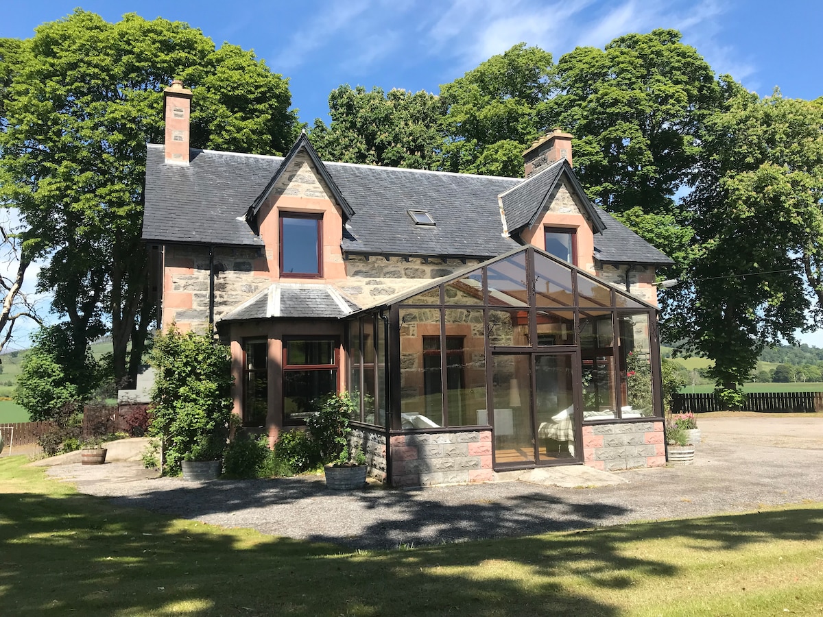 A charming two-story house is surrounded by green trees and open grounds. The front features a glass conservatory that allows for natural light, with a mix of stone and rendered walls. A gravel driveway leads to the entrance, complementing the tranquil outdoor setting.