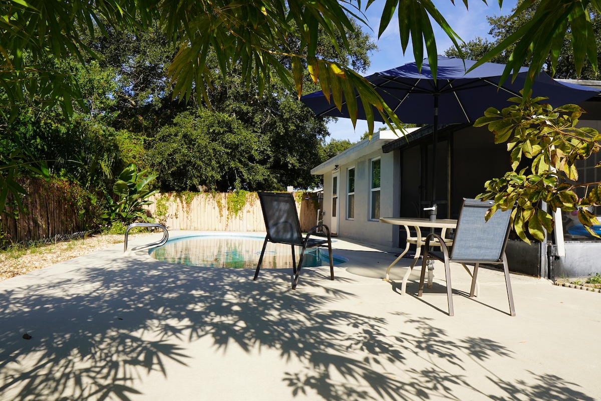 A private outdoor space features a clear swimming pool surrounded by concrete. Several chairs and a table are positioned near a large umbrella, providing shade. Lush green foliage frames the area, while the house with multiple windows is visible in the background.
