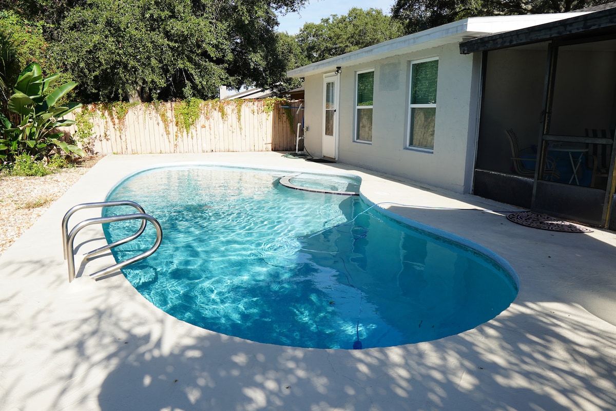 A refreshing pool is set in a private outdoor area, surrounded by greenery. The pool features a gently curving shape with a ladder for easy access. The space includes a shaded seating area nearby, offering a perfect spot to relax.