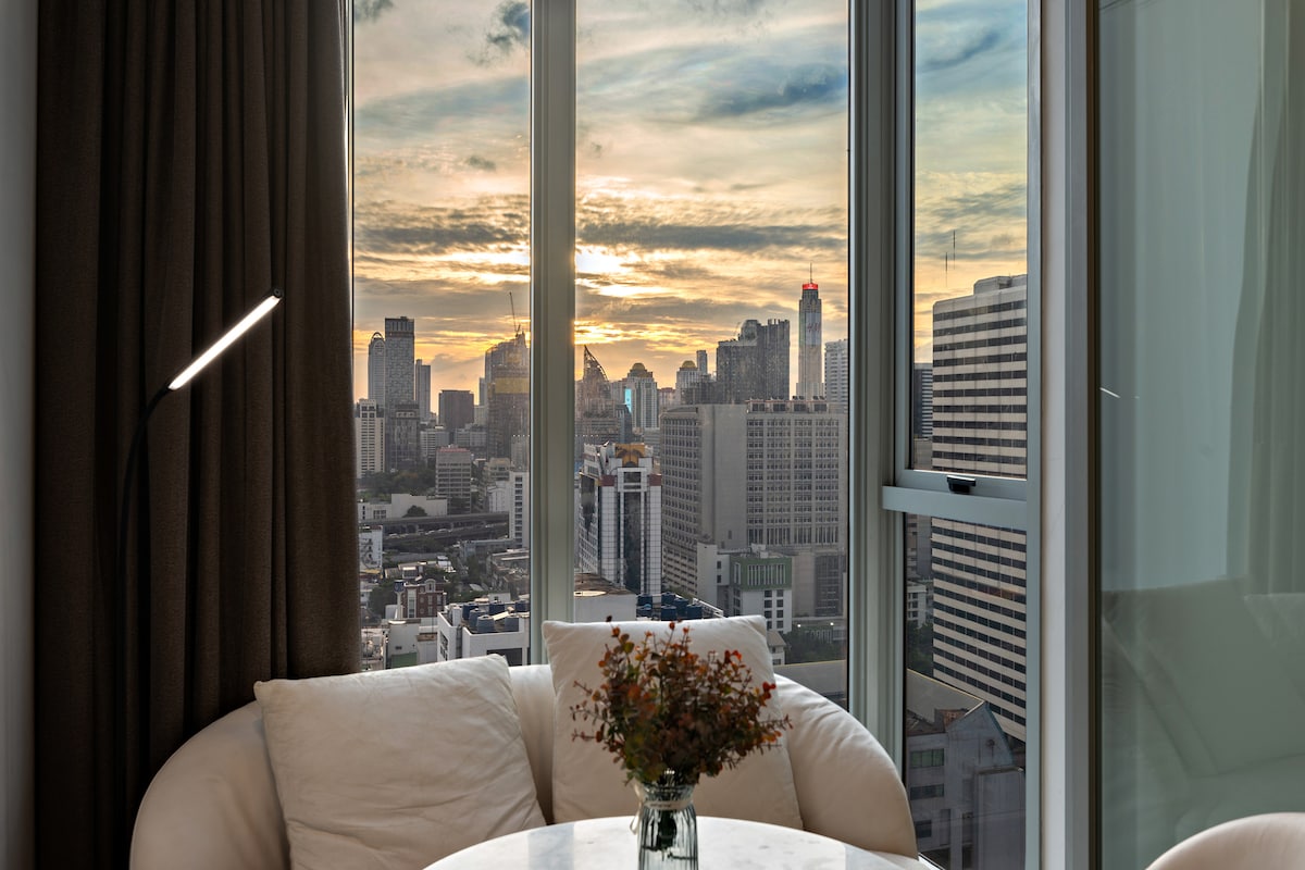 A cozy seating area features a round marble table accompanied by a plush armchair. Large windows offer a panoramic view of the city skyline at sunset, with golden hues illuminating the scene. A small vase with fresh flowers is placed on the table.