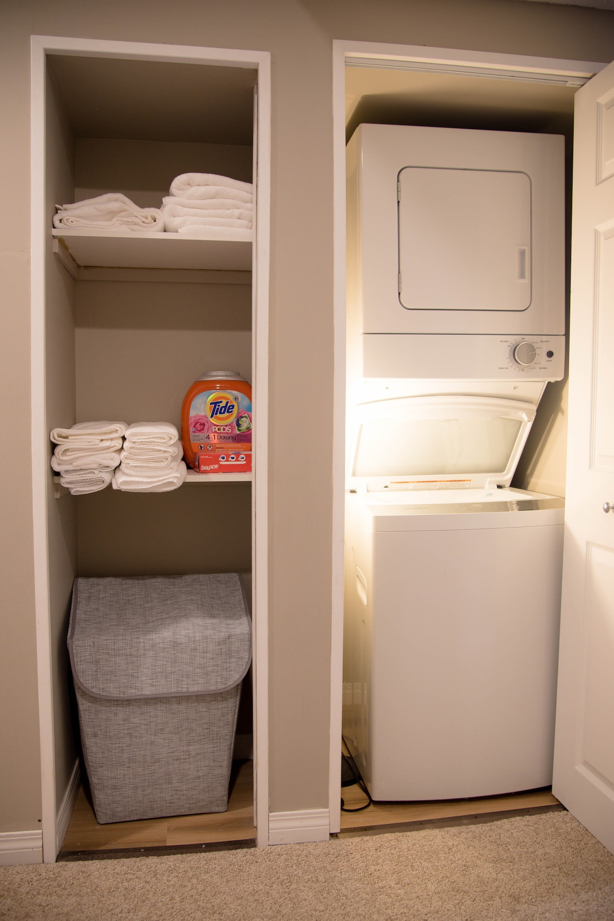 A neatly organized laundry area features a stackable washer and dryer. A shelf holds several neatly folded towels, and a laundry basket is positioned beneath. Detergent is clearly displayed on the shelf, ensuring convenience for guests during their stay.