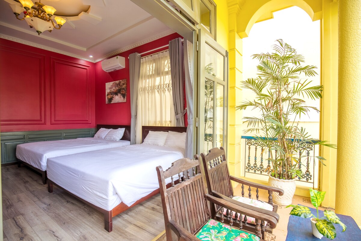 A bright room is featured with two spacious beds, complemented by a vibrant red accent wall. Natural light pours in through large windows, while a small balcony with wooden chairs overlooks a potted plant. The overall ambiance is airy and inviting.