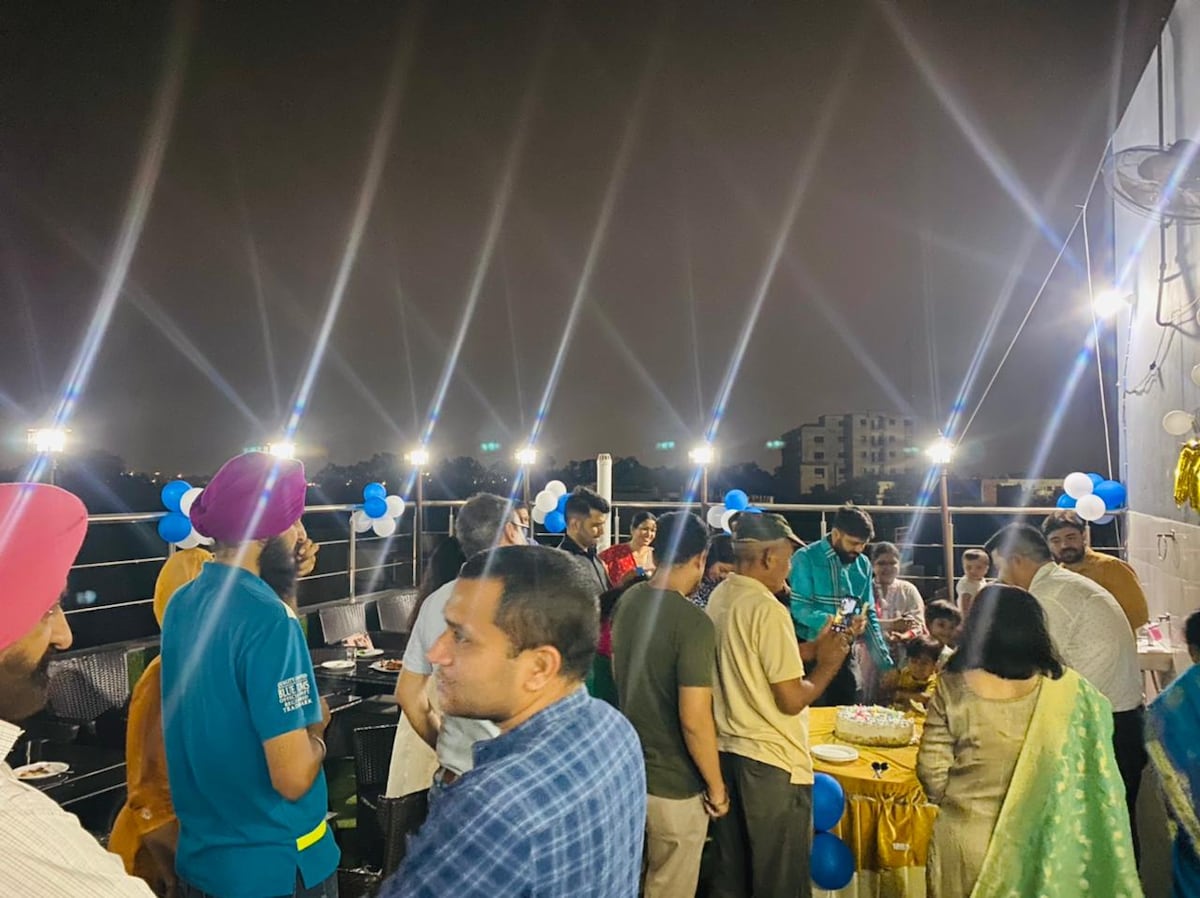 A lively gathering is taking place on an outdoor terrace, with guests mingling under strings of bright lights. Balloons are seen in shades of blue, and a buffet table with a refreshing spread is positioned in the background. The city skyline is faintly visible at night.