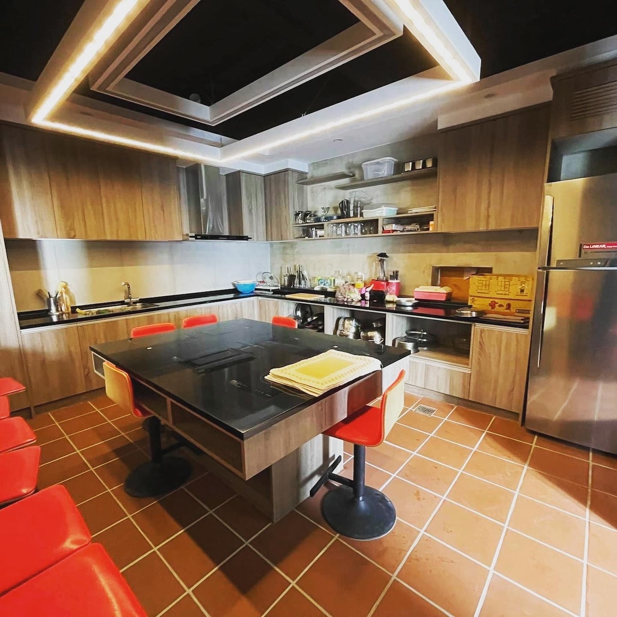 The kitchen features modern wooden cabinetry and sleek black countertops. An island with bar stools offers additional seating. Shelves hold various kitchen items and utensils, while stainless steel appliances are positioned along the wall, providing a functional cooking space.