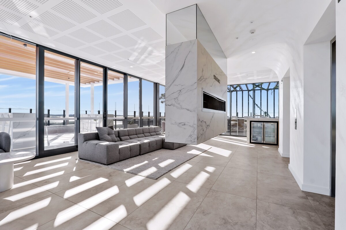 An open, airy living space is highlighted by large windows that invite natural light. A minimalist gray sofa sits on a light-colored floor, complemented by a marble feature wall. Stylish furnishings enhance the modern design, creating a connected feel to the outdoor views.