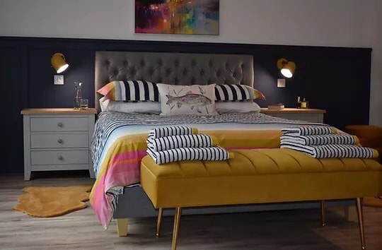 A modern bedroom features a spacious bed with a tufted gray headboard, complemented by coordinating striped linens and decorative pillows. A yellow upholstered bench sits at the foot of the bed. Two bedside lamps provide warm lighting, and a contemporary artwork piece hangs above the bed.