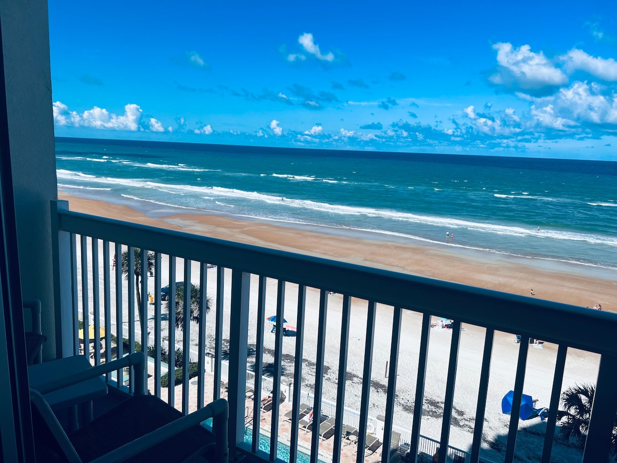 The private balcony offers an unobstructed view of the Atlantic Ocean, showcasing expansive sandy beaches and the gently rolling waves. Clear blue skies and fluffy white clouds enhance the serene coastal landscape. A glimpse of beachgoers can be seen below, enjoying the shoreline.