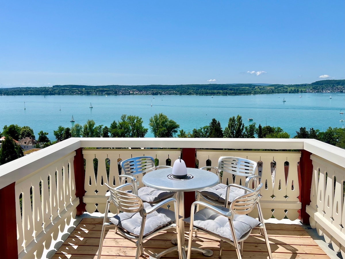 A scenic balcony features a round table and two chairs, providing a space to enjoy panoramic views of the Bodensee. The calm water reflects the clear blue sky, while lush greenery lines the distant shores, contributing to the overall serene atmosphere.