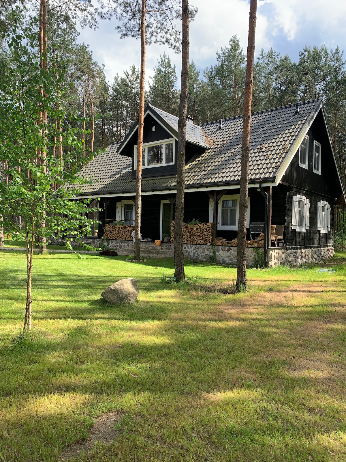 A traditional wooden house reflects charming architectural details, set amidst a lush green landscape. The property features a sloped roof and a stone foundation, with logs stacked by the side. Tall trees provide shade, enhancing the tranquil environment.