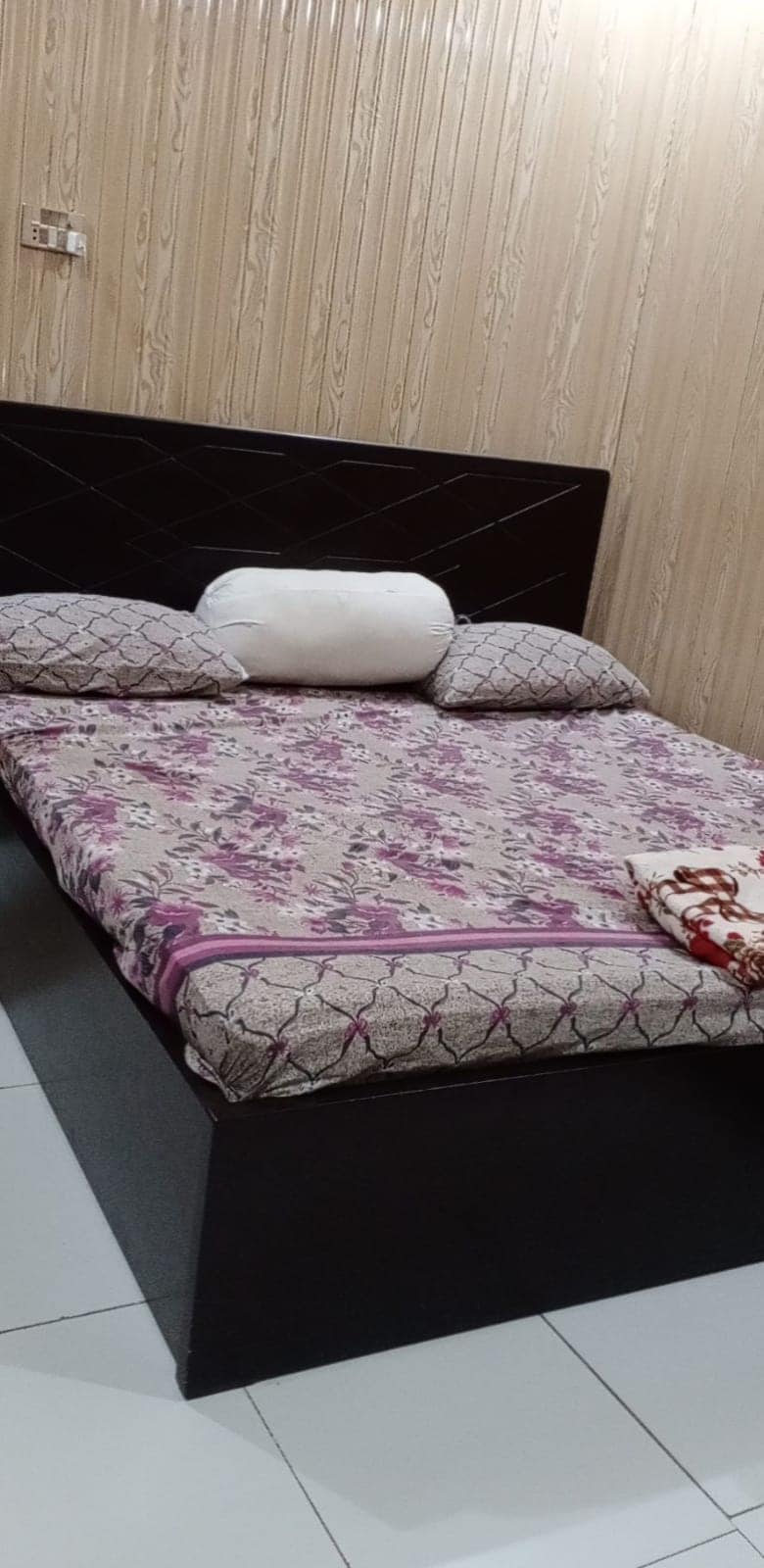 A cozy sleeping area features a bed adorned with a floral-patterned blanket and two decorative pillows. The headboard presents a sleek design, and the room's walls are clad in simple wood paneling, complementing the tiled floor.