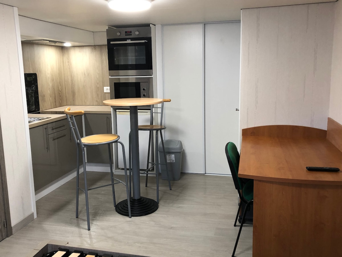 A modern kitchenette is presented with sleek cabinets and a built-in oven, accompanied by an induction cooktop. A tall round table with two stools is visible, along with a small writing desk and a green chair. The open design offers a spacious and functional layout.