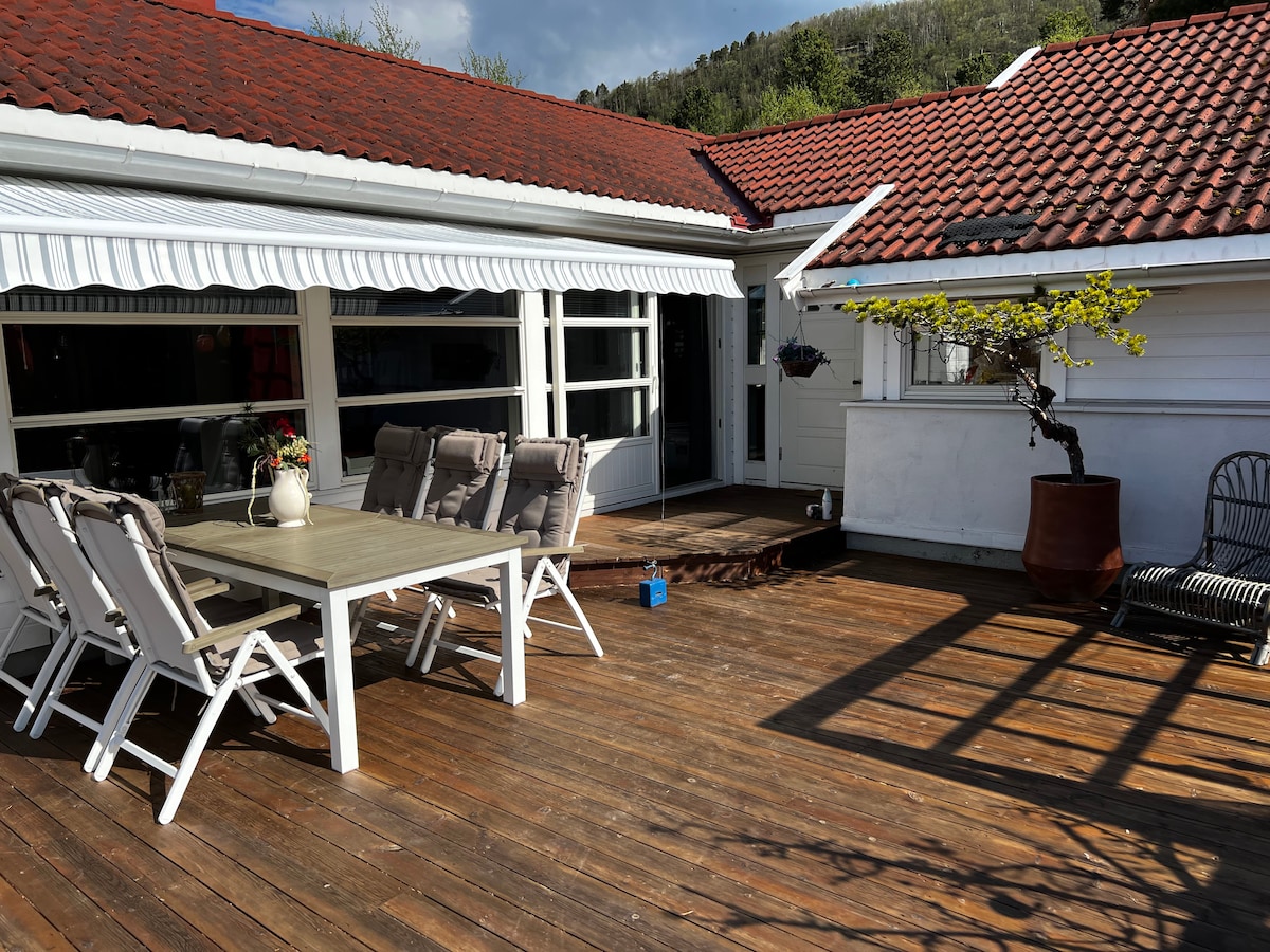 An outdoor seating area features a spacious wooden deck with a large table and six chairs. A shaded area is provided by an awning, and a decorative plant sits in a large pot. Natural light highlights the wood grains of the deck.