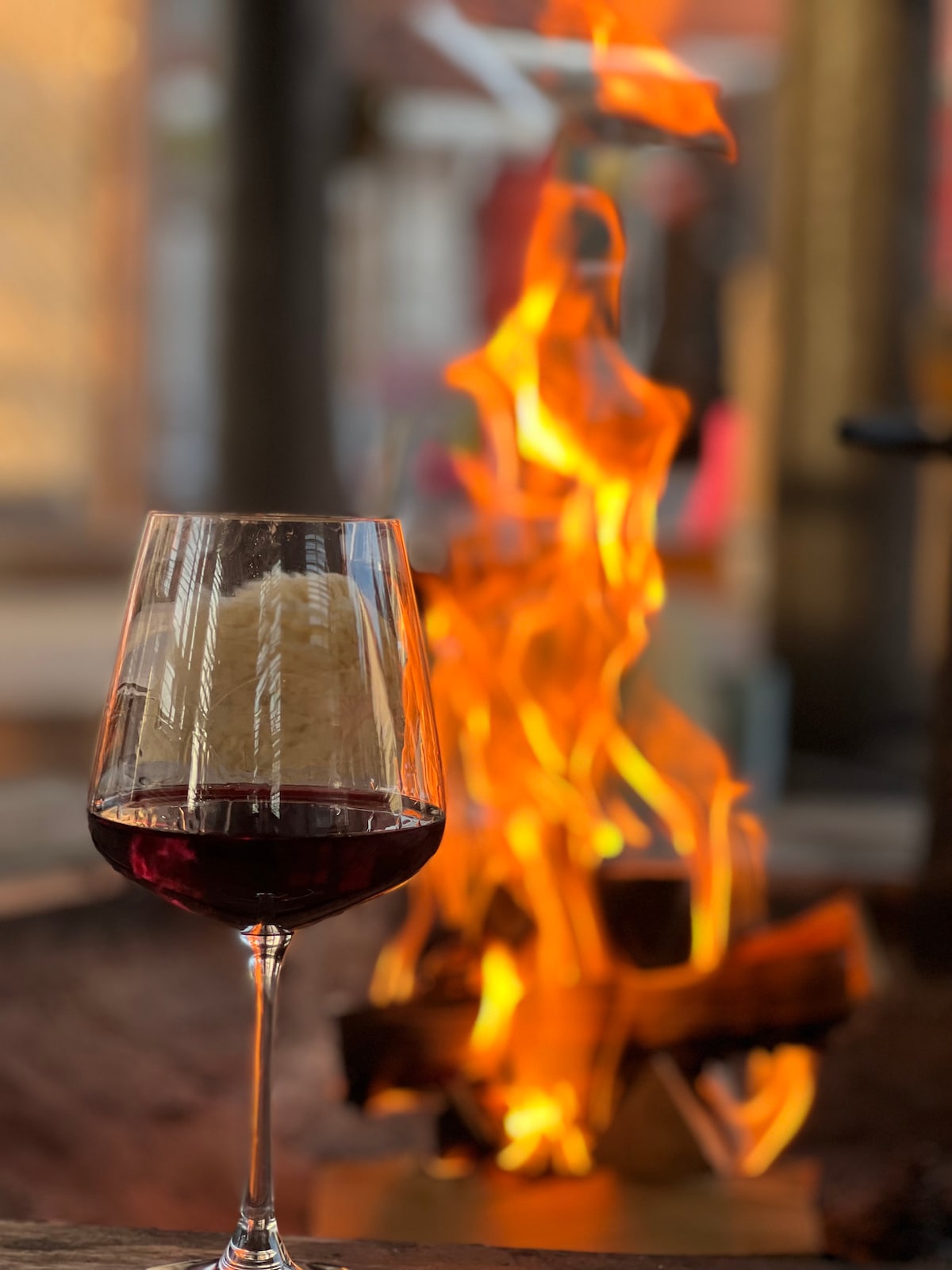 A glass filled with deep red wine is placed in front of a vibrant flame. The warm glow from the fire creates a cozy atmosphere, highlighting the wine's rich color against the backdrop of glowing embers.