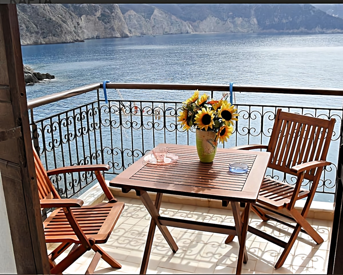 A balcony overlooking a calm sea features a wooden table surrounded by two folding chairs. A vibrant flower pot with sunflowers adds color to the setting. The railing, designed with intricate patterns, frames the stunning water view and distant cliffs.