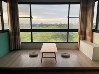 A cozy seating area features a low wooden table with two cushions on a spacious wooden floor. Large windows provide a sweeping view of the lush green landscape outside, while soft natural light fills the room.