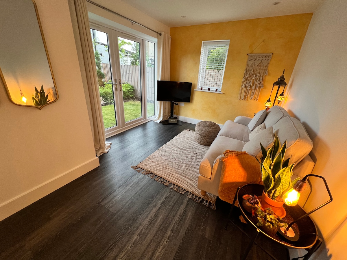 A lounge area is presented with natural light, featuring a comfortable sofa and a flat-screen TV. A rug lies beneath a circular table adorned with decorative items. A mirror reflects the space, while double doors open to an outdoor view, creating a sense of openness.