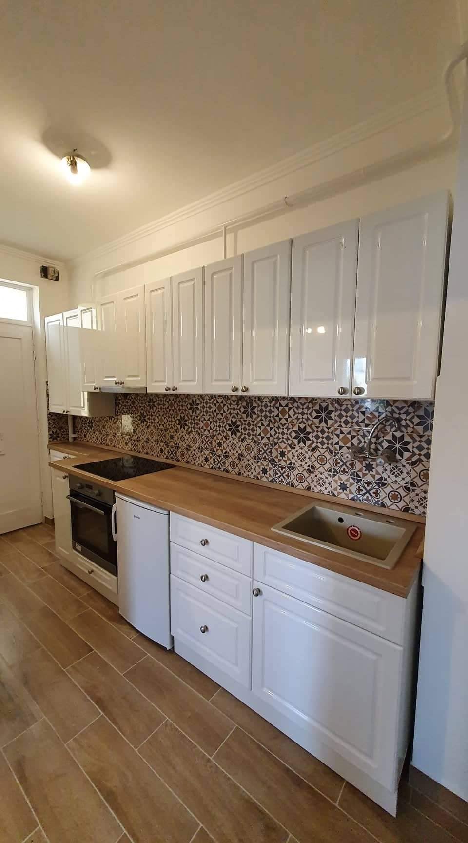 A modern kitchen features sleek, white cabinetry complemented by a decorative backsplash. Stainless steel appliances, including an oven and refrigerator, are included. A compact sink is integrated into the wooden countertop, adding functionality to the space, while natural light streams in through a nearby window.