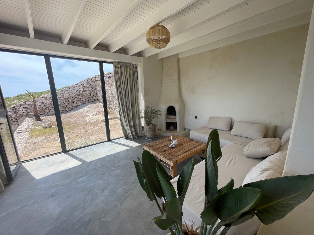 A spacious living area features a comfortable seating arrangement with cushioned sofas. Natural light floods the space through large glass doors, highlighting the minimalist design. A wooden coffee table sits in the center, accompanied by indoor plants set against an earthy backdrop.