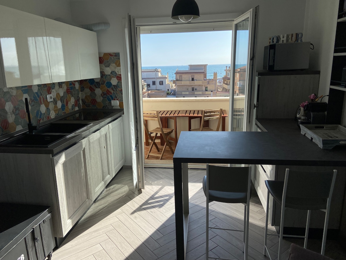 A modern kitchen area is visible with sleek cabinetry and a colorful backsplash. Large glass doors open to a balcony, showcasing sea views. A dining table with chairs is positioned in the space, allowing for a bright, airy atmosphere filled with natural light.