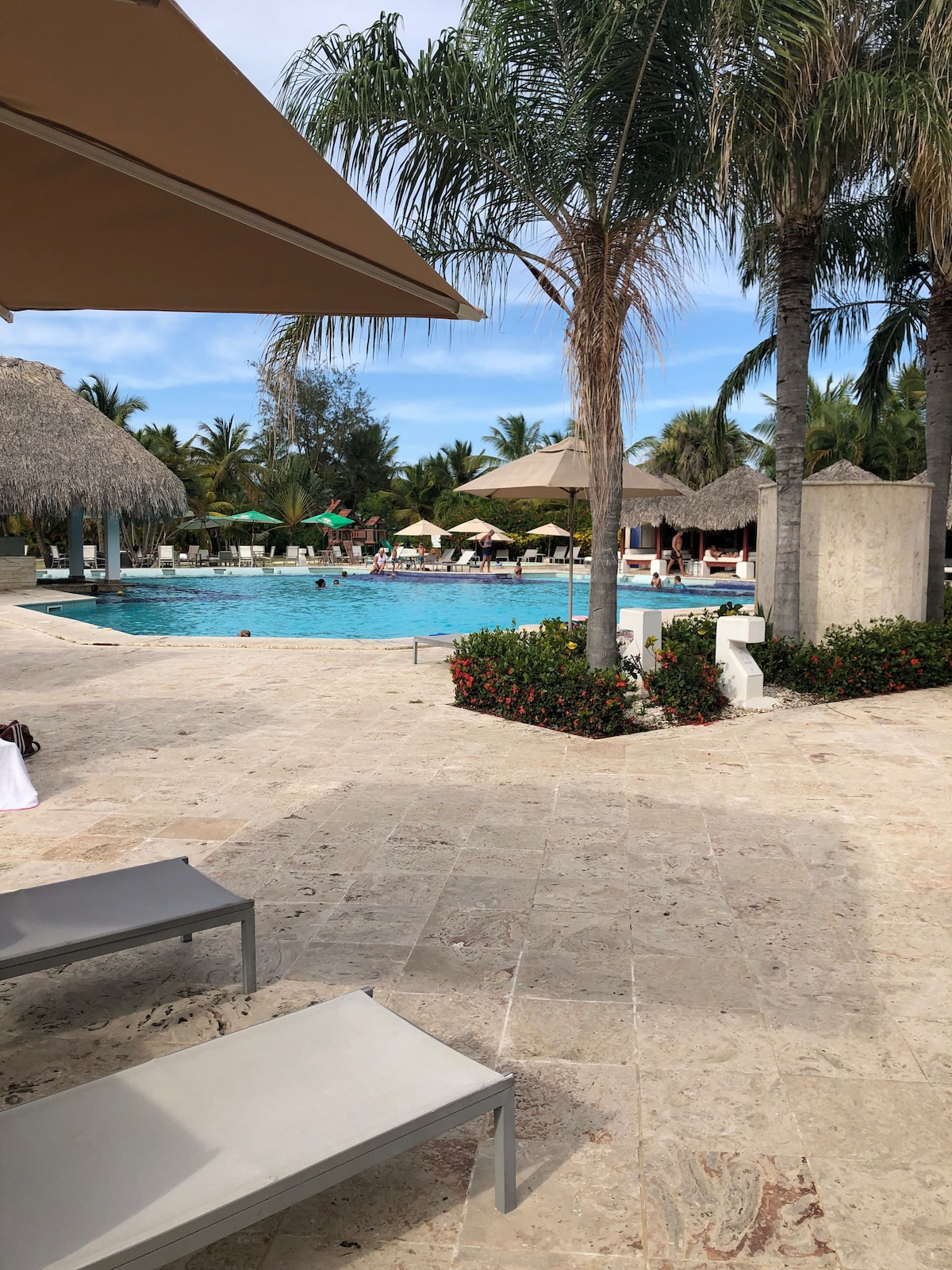A spacious pool area is surrounded by tropical palm trees and thatched-roof umbrellas. Loungers are positioned on a tiled patio, offering views of the inviting pool and lush landscaping. Guests can be seen relaxing by the water, enhancing the serene environment.