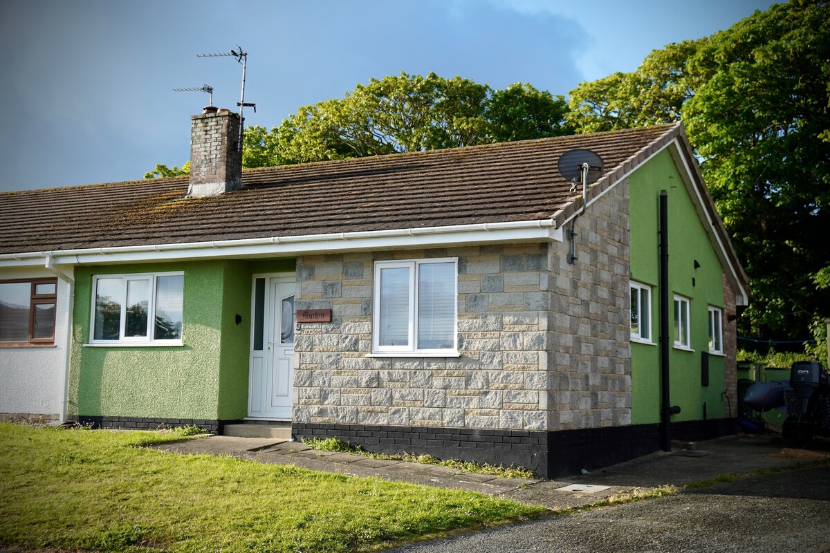 The exterior of a single-story bungalow is shown, featuring a mix of stone and green siding. A well-kept lawn surrounds the entrance, which has a white door and windows that provide natural light. Trees are visible in the background, adding to the serene setting.