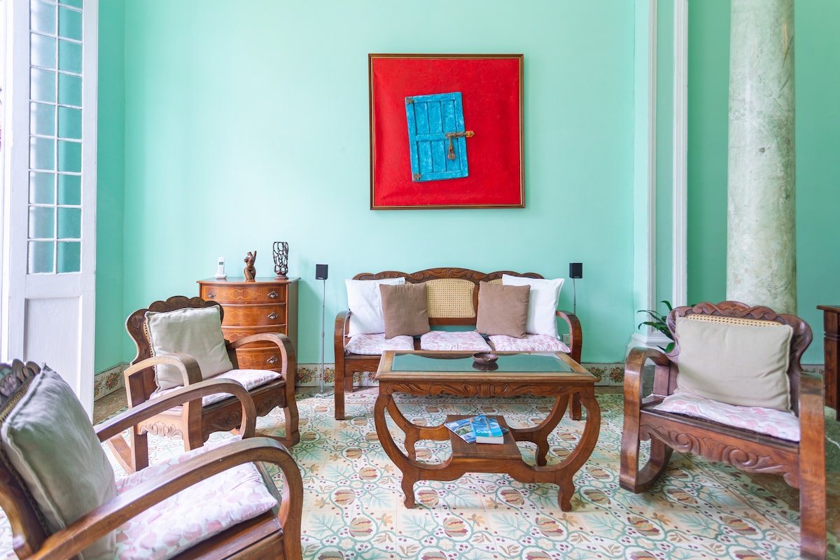 A bright living area features light green walls and colorful artwork. Wooden furniture, including a coffee table and several armchairs, offers seating around the space. A patterned tile floor complements the room's decor, while large windows provide natural light.