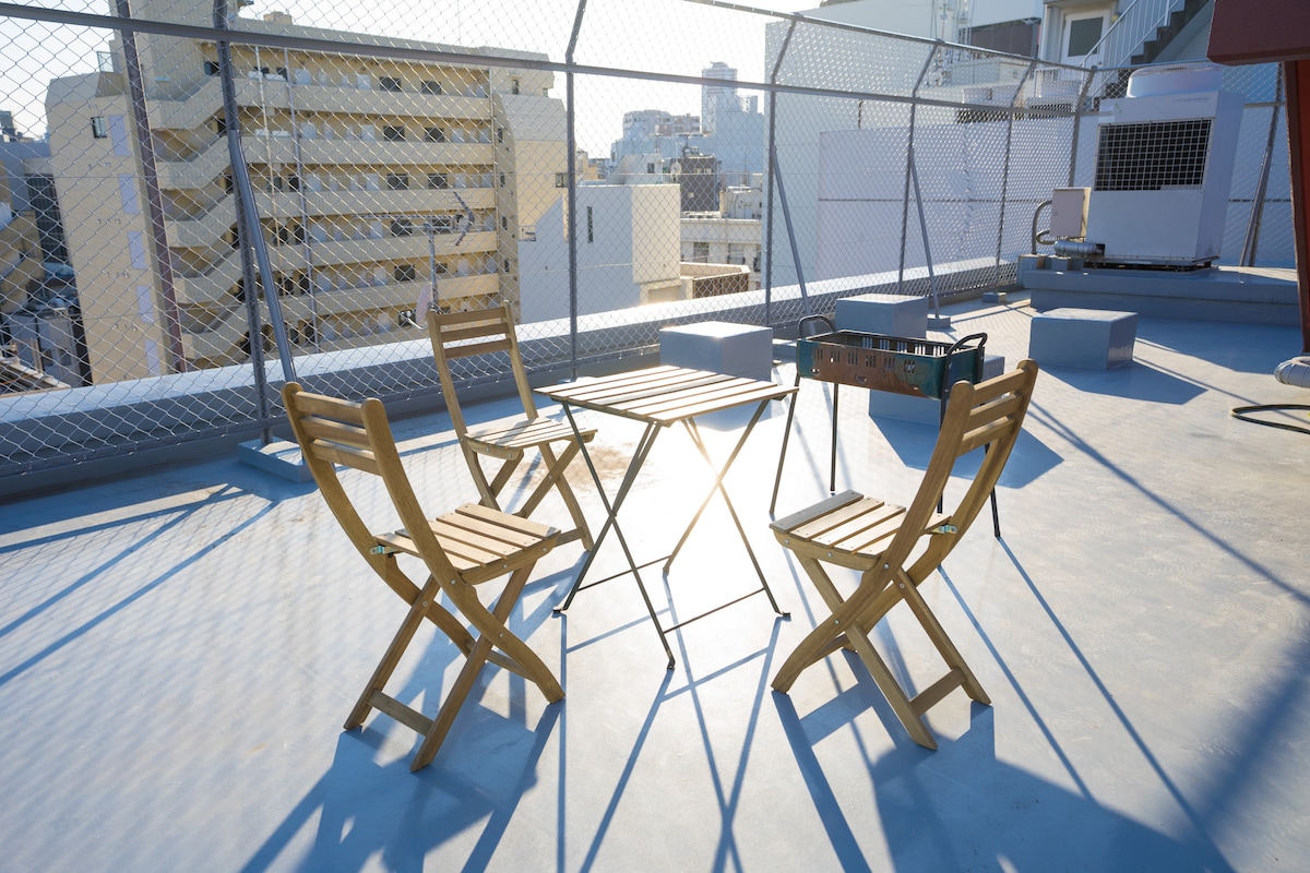 A rooftop space is equipped with a small table and four wooden chairs. The area is surrounded by a safety fence, offering a view of the city skyline. Natural light creates reflections on the surface, enhancing the spacious feel.