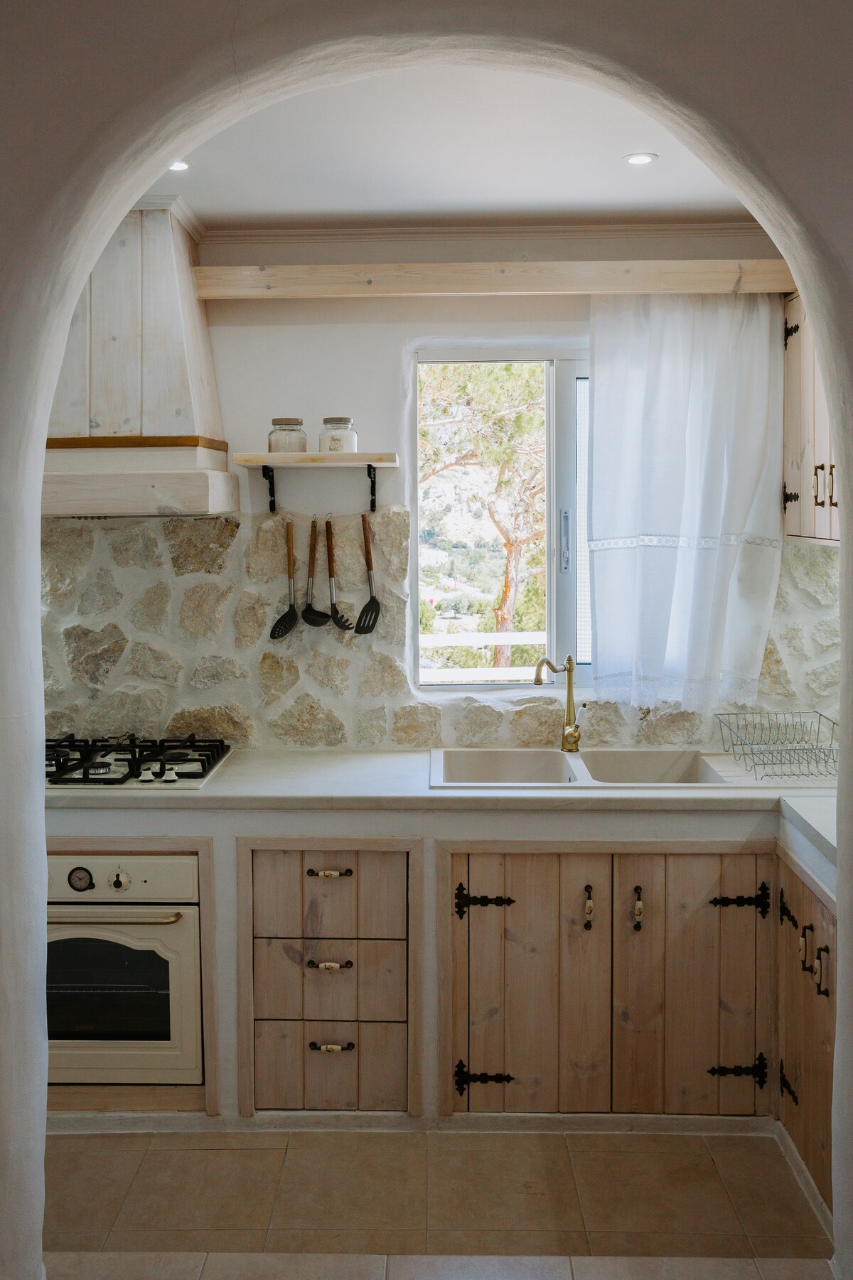 A modern kitchen features natural stone walls and wooden cabinetry, offering an open window that allows light to stream in. A stove, oven, and sink are conveniently arranged, with cooking utensils displayed neatly.