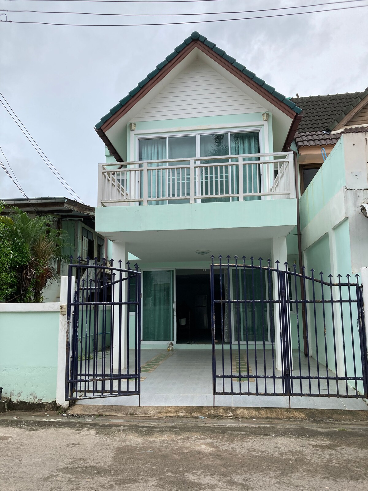 A two-story house is presented with a sloped roof and a light green faรงade. A gated entrance leads to a spacious patio area, framed by glass doors that offer a clear view of the interior. The surrounding area features greenery and nearby structures.