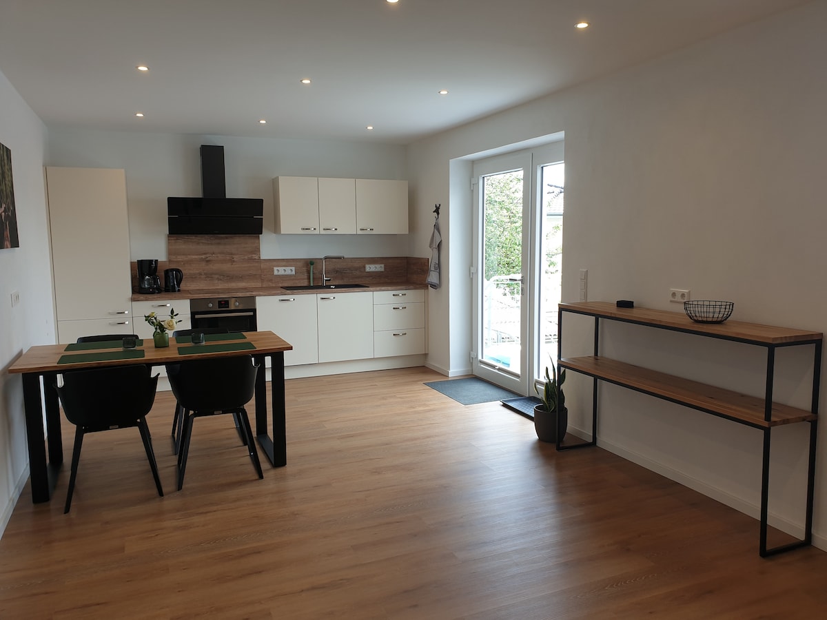A modern kitchen space is shown, featuring sleek white cabinetry and wooden accents. A dining table with black chairs is positioned in the center, while a side table displays plants. Natural light enters through a large window, enhancing the bright and functional layout.