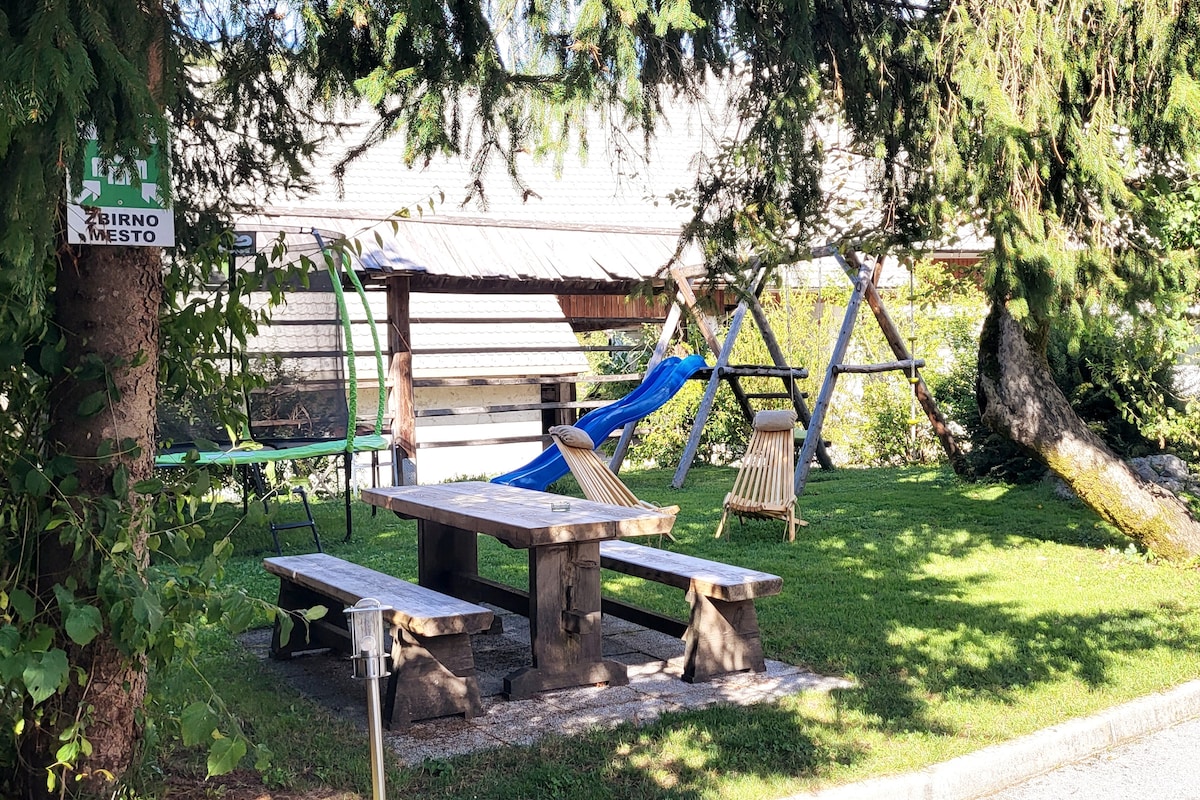 A garden area features a wooden table surrounded by benches, offering a space for dining. Nearby, a swing set and a slide are visible, along with a hammock and a small structure with a roof. Lush greenery provides a natural backdrop.