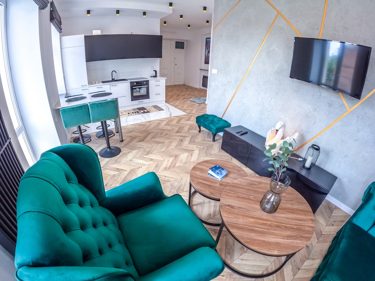 A modern living area features plush green seating arranged around a round wooden coffee table. The space includes a sleek kitchen visible in the background, with dark cabinetry and stainless steel appliances. A television is mounted on the wall, complementing the contemporary design.