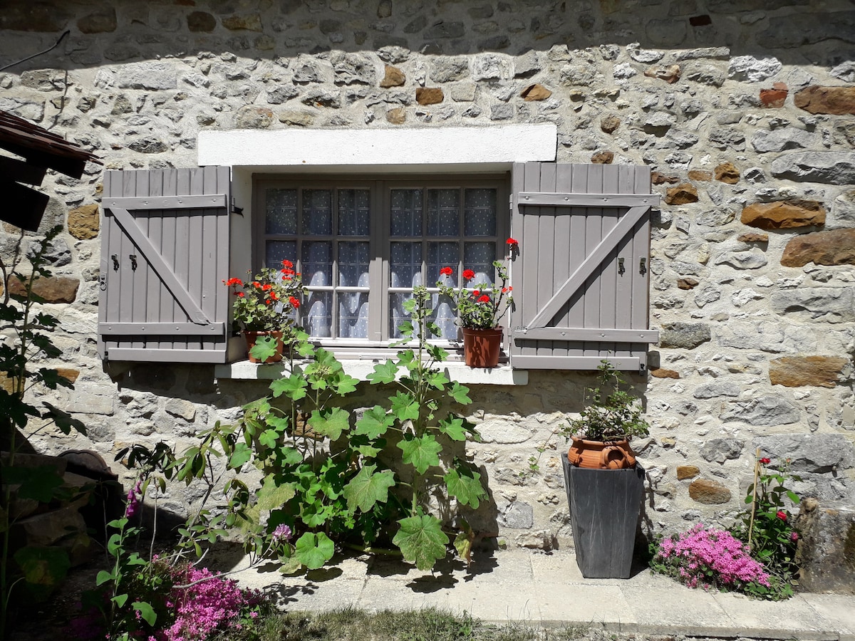 A charming stone wall is enhanced by shuttered windows, which feature flower boxes filled with vibrant blooms. Lush green plants emerge from the ground below, while colorful flowers add a touch of brightness, creating a welcoming appearance to the outdoor space.