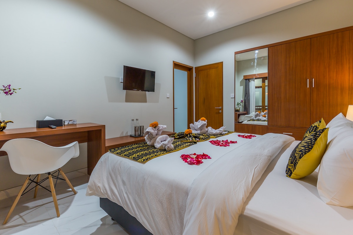 A spacious bedroom is decorated with floral arrangements on the bed. An LCD TV is mounted on the wall across from the bed. A desk and chair are positioned to the side, while wooden cabinets provide additional storage. A mirrored wardrobe reflects the room's calming ambiance.