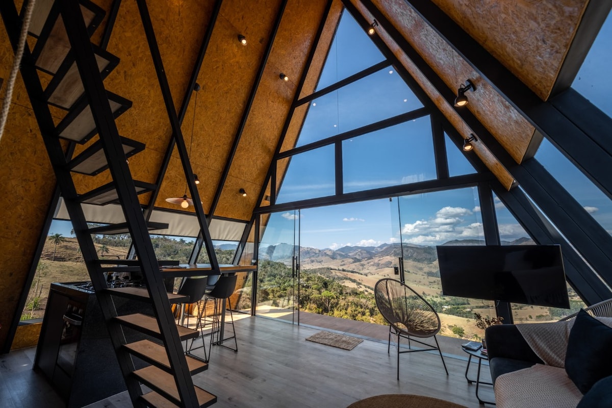 The interior showcases a spacious, open-plan living area with large glass windows offering panoramic mountain views. A modern staircase ascends, complemented by a dining table and chairs, and a comfortable seating area with a television. The unique A-frame architecture enhances the natural light throughout the space.