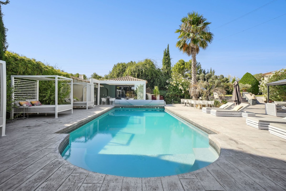 A serene outdoor area features a clear swimming pool surrounded by wooden decking. Lush greenery borders the space, complemented by scattered lounge chairs and shaded seating areas. A palm tree adds a tropical touch to the calm atmosphere.